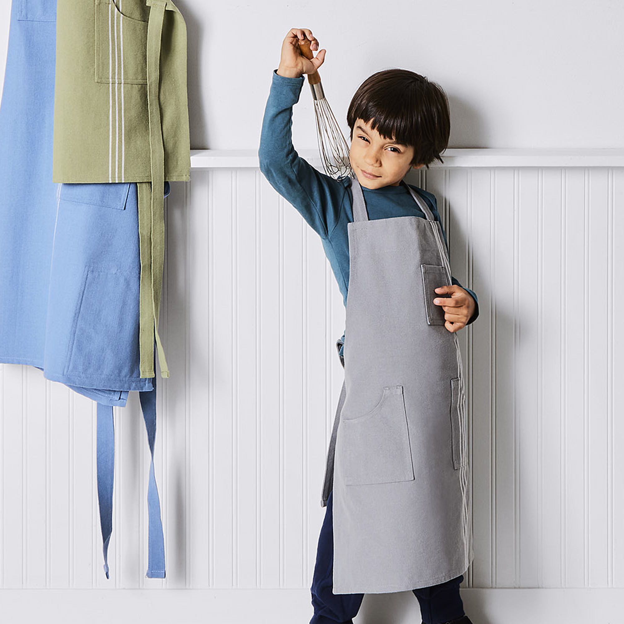 A young boy cheekily showing off a gray apron with a whisk in his hand. Aprons in rose, soft blue, and sage hang behind him on a wall.