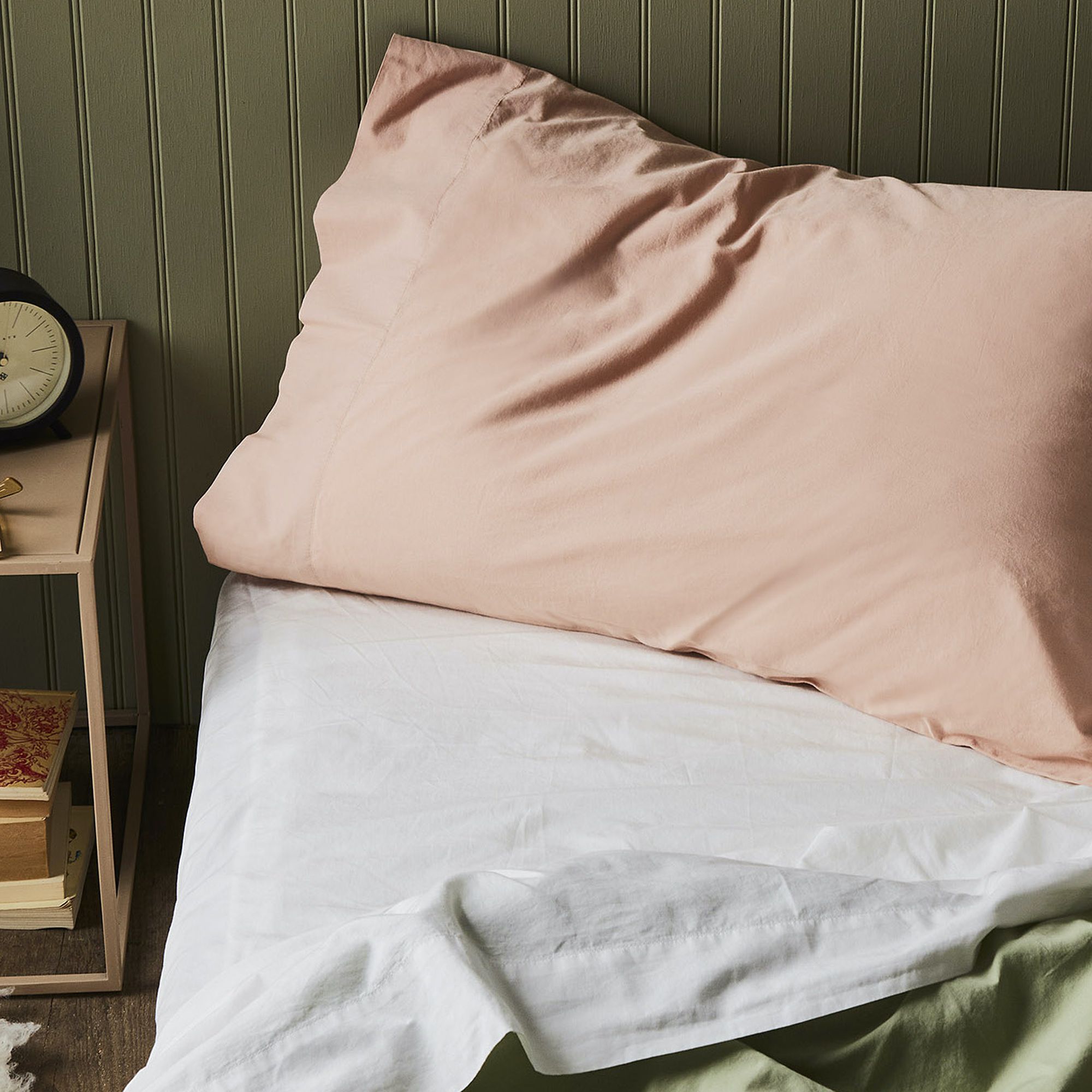 A bed with pink pillows, against a dark green wall, a small nightstand.